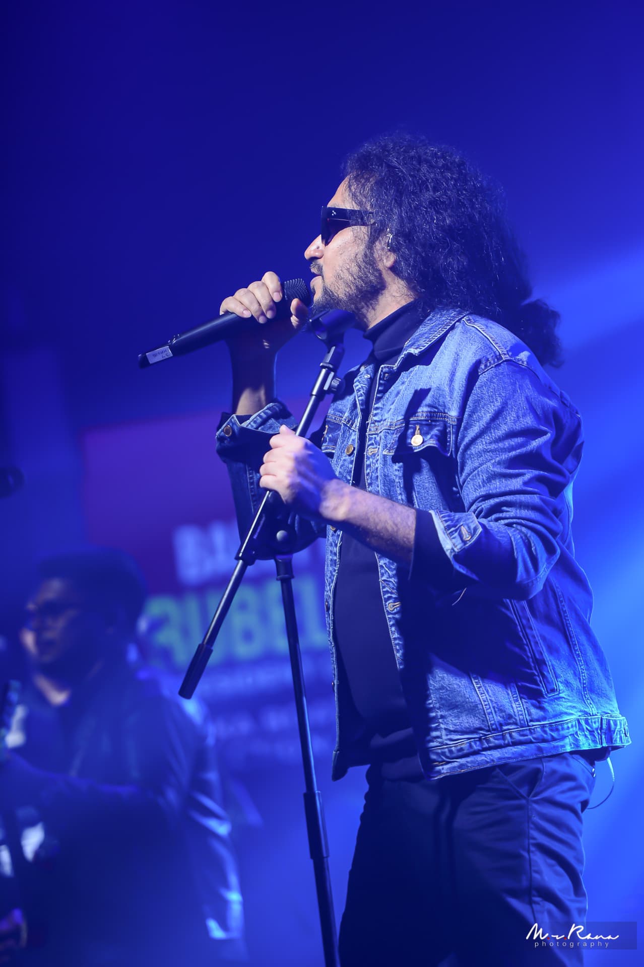 Muttaque Hasib performing live on stage in blue light, singing into a handheld microphone
